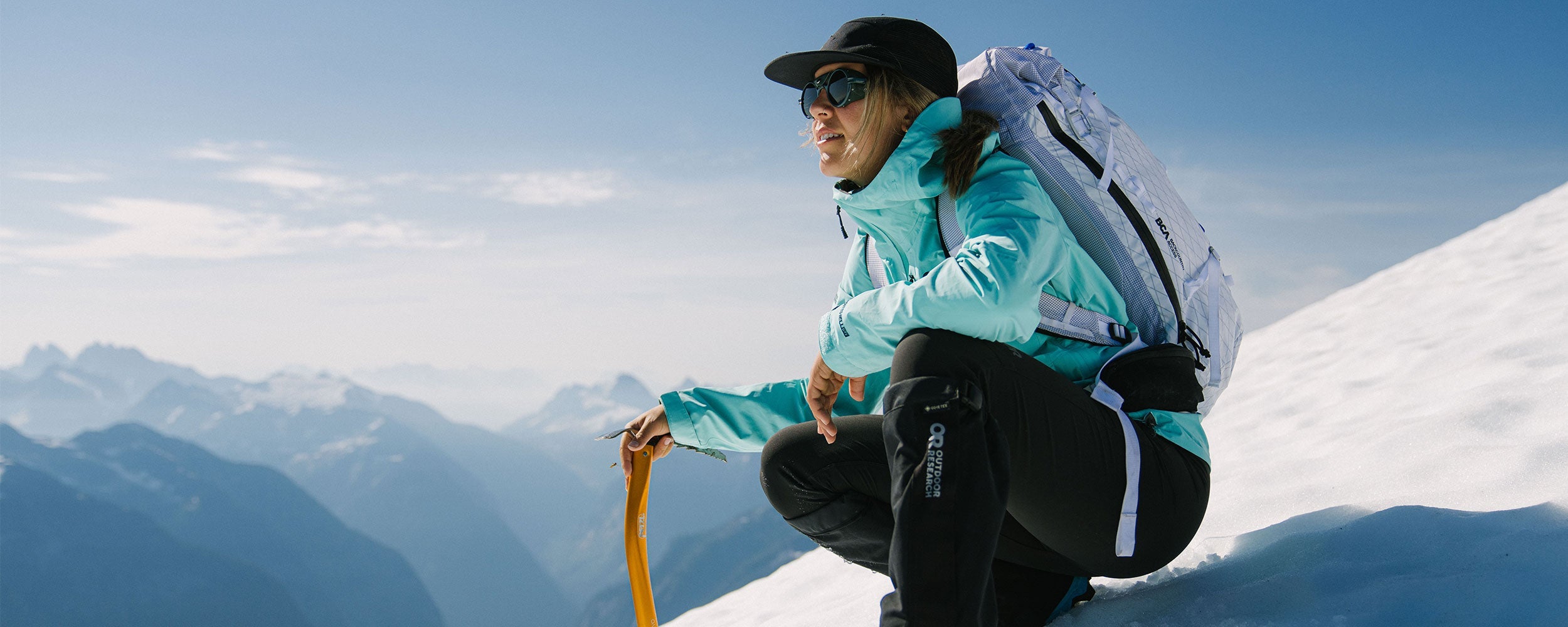 Sun beams down on a woman crouching during her hike while wearing Outdoor Research Aspire 3L Jacket in maldives/blue.