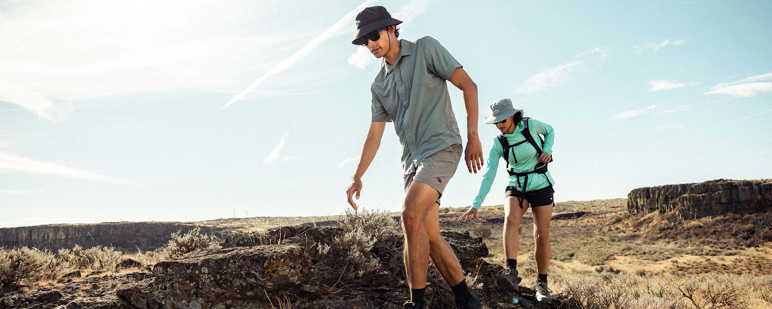 Two friends balance on rock as they hike in the sun wearing Outdoor Research sportswear. 