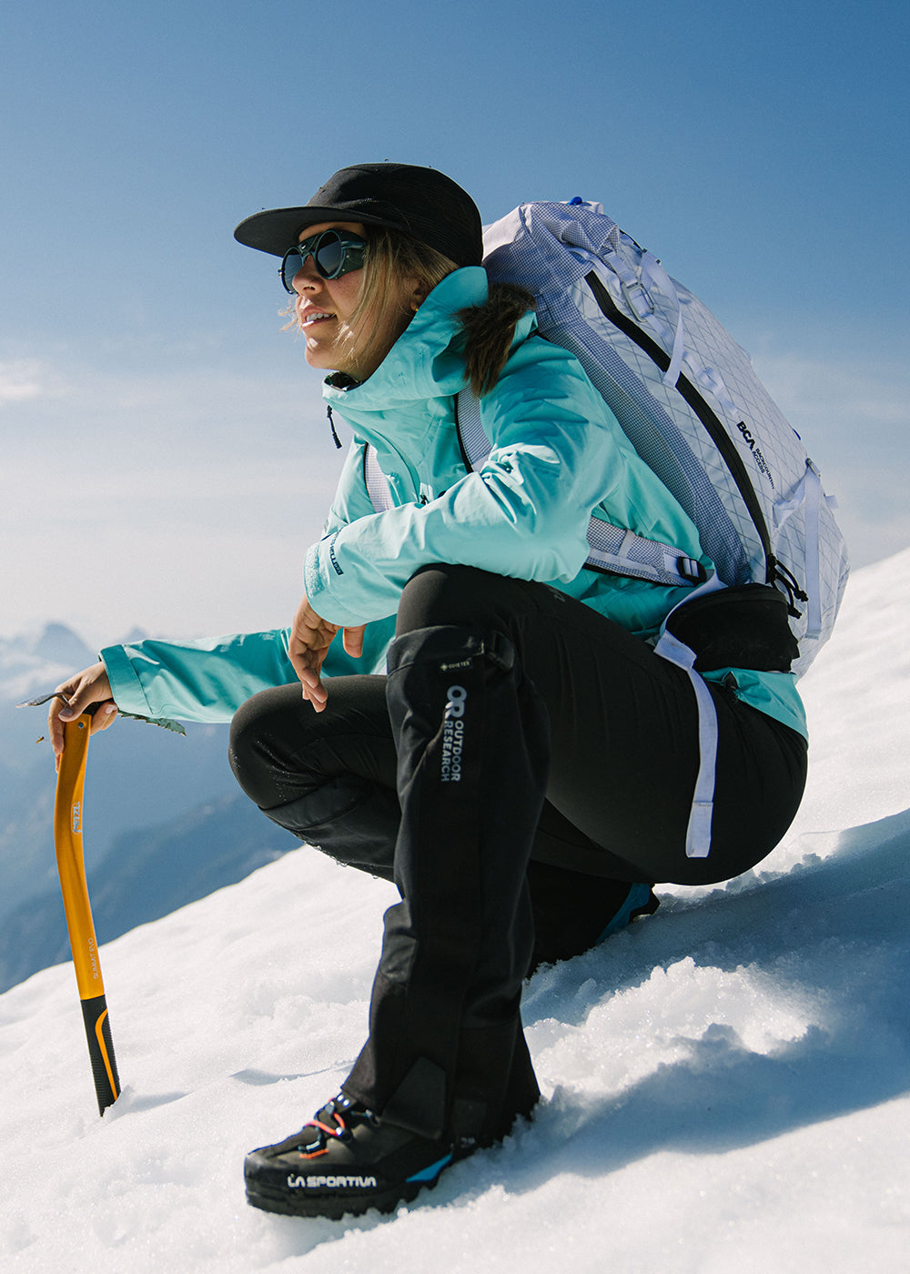 Sun beams down on a woman crouching during her hike while wearing Outdoor Research Aspire 3L Jacket in maldives/blue.